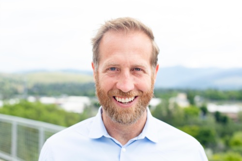 Headshot of Jake Harriman, a white man with a salt and pepper beard standing in front of scenic mountains