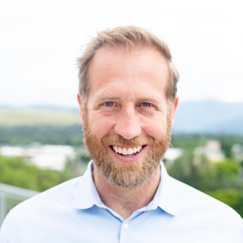 Headshot of Jake Harriman, a white man with a salt and pepper beard standing in front of scenic mountains