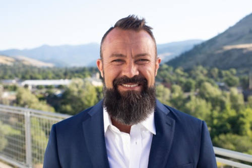 headshot of Shon Westfall, a white man with a beard wearing a blue suit, with mountains in the background