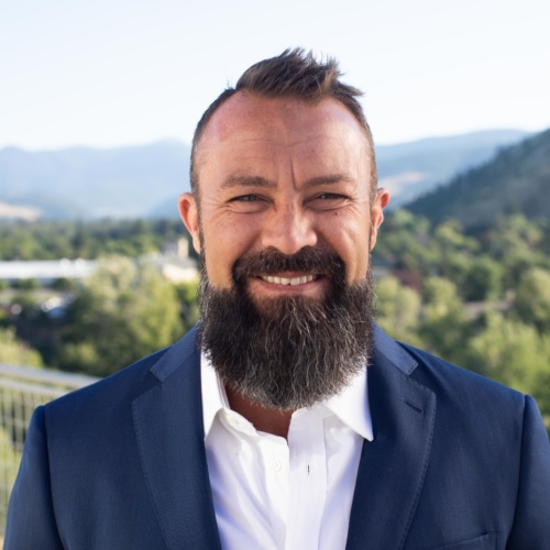 headshot of Shon Westfall, a white man with a beard wearing a blue suit, with mountains in the background