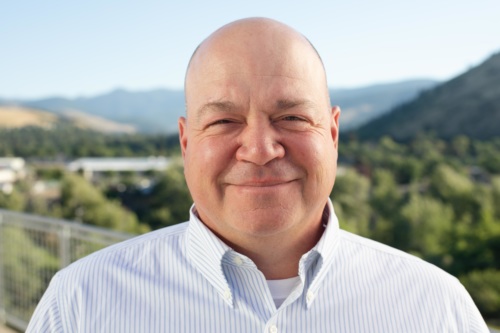headshot of Brandon Newton, a bald white man wearing a button up blue shirt, and mountains in the background