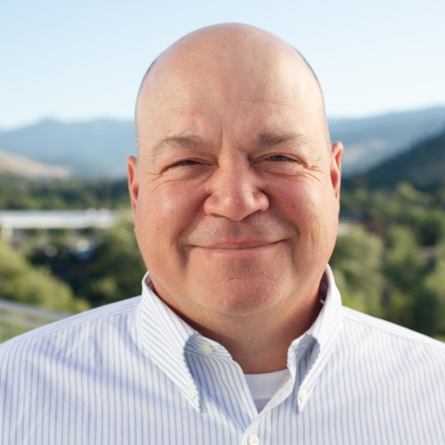headshot of Brandon Newton, a bald white man wearing a button up blue shirt, and mountains in the background