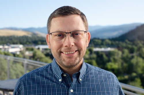 headshot of Michael Greenwood, a white man with glasses wearing a blue button up shirt, with mountains in the background