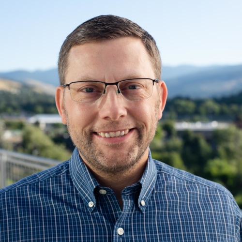 headshot of Michael Greenwood, a white man with glasses wearing a blue button up shirt, with mountains in the background
