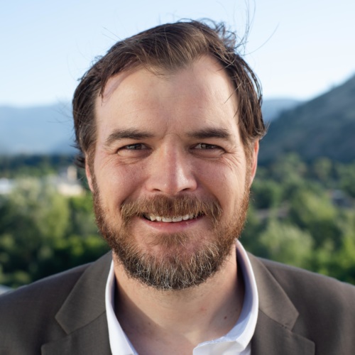 headshot of Ryan Woolf, a white man wearing a grey suite, with mountains in the background