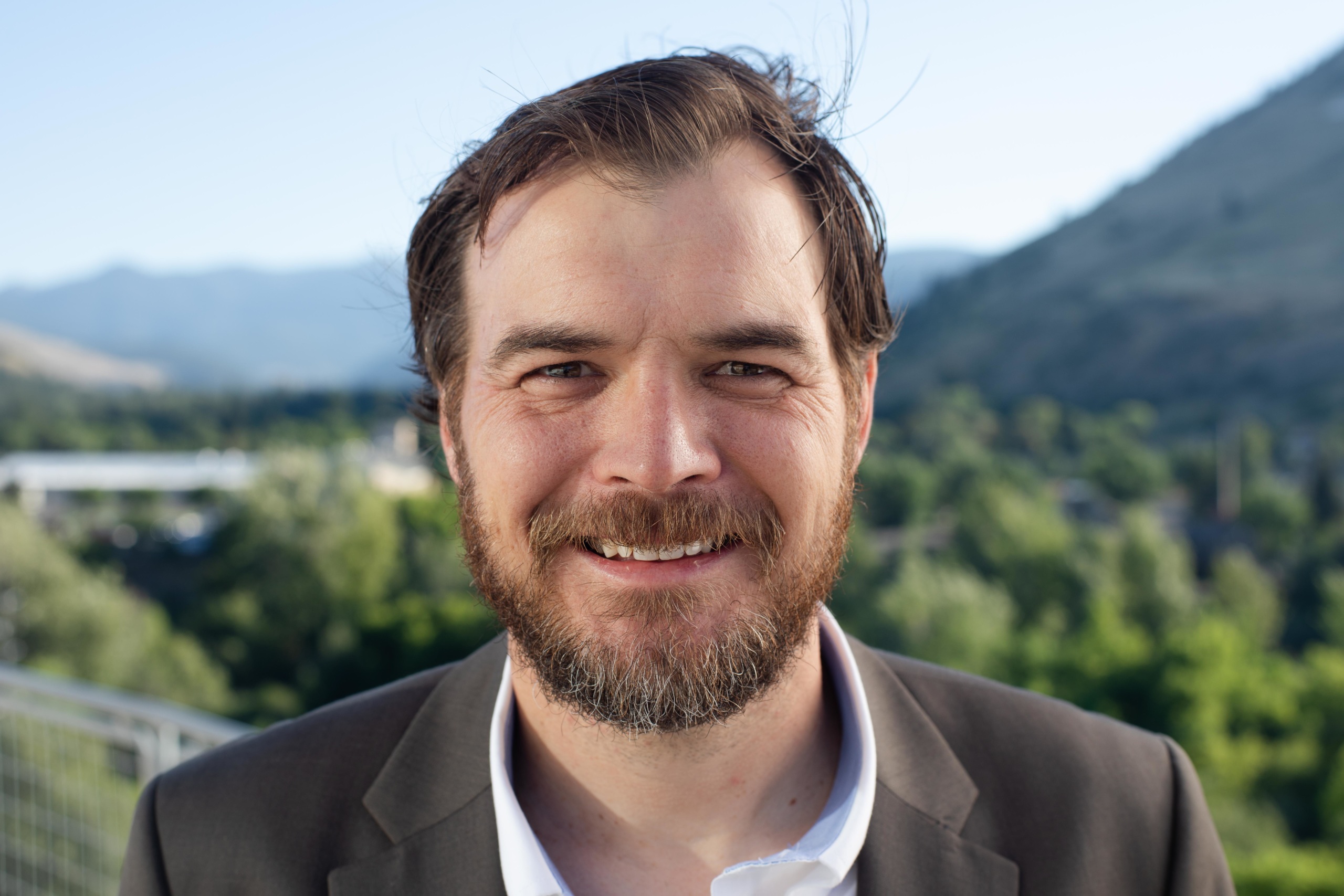 headshot of Ryan Woolf, a white man wearing a grey suite, with mountains in the background