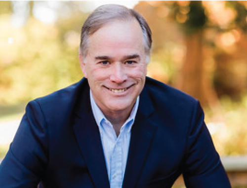 headshot of trey lackey, an older white man dressed in a blue jacket and blue button up shirt with foliage in the background.