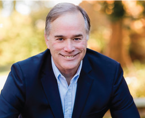 headshot of trey lackey, an older white man dressed in a blue jacket and blue button up shirt with foliage in the background.