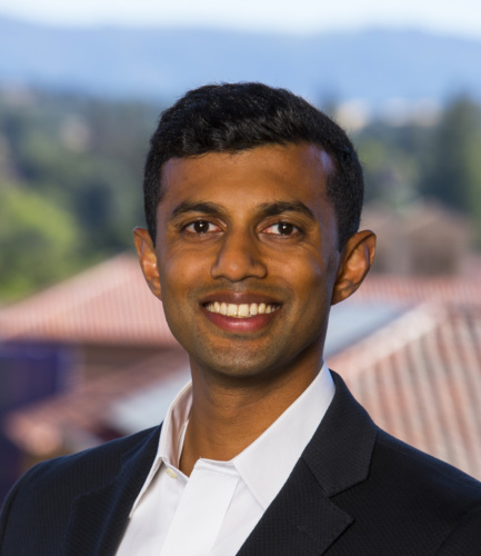 Indian American man in black jacket and white shirt headshot with nature in background.