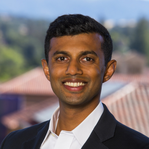 Indian American man in black jacket and white shirt headshot with nature in background.