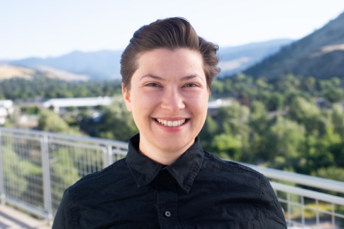 headshot of Victoria Rametta, white nonbinary person with short brown hair, in a black button up shirt with mountains in the background.