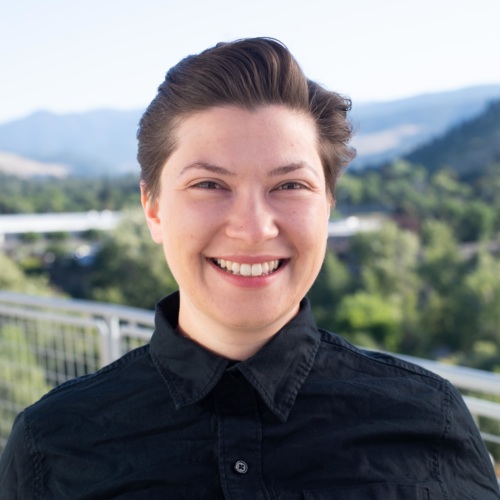 headshot of Victoria Rametta, white nonbinary person with short brown hair, in a black button up shirt with mountains in the background.