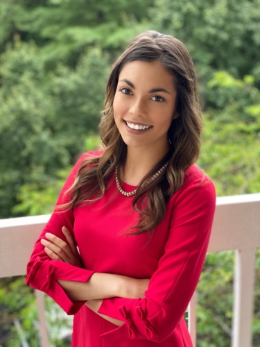 Headshot of a lightskinned woman with long dark chair wearing a red long sleeved shirt with trees in the background.