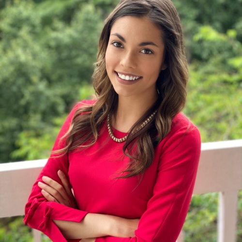 Headshot of a lightskinned woman with long dark chair wearing a red long sleeved shirt with trees in the background.