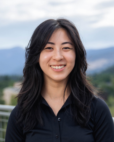Headshot of Maina Chen, an Asian woman with long dark wavy hair in a black dress shirt with mountains in the background.