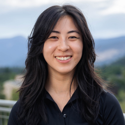 Headshot of Maina Chen, an Asian woman with long dark wavy hair in a black dress shirt with mountains in the background.