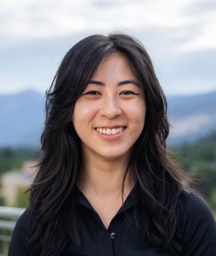 Headshot of Maina Chen, an Asian woman with long dark wavy hair in a black dress shirt with mountains in the background.