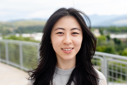 headshot of Maina Chen, an Asian woman with long dark hair in a grey and beige sweater with mountains in the background.