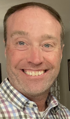 Headshot of a white, middle-aged man smiling in a checkered button up shirt