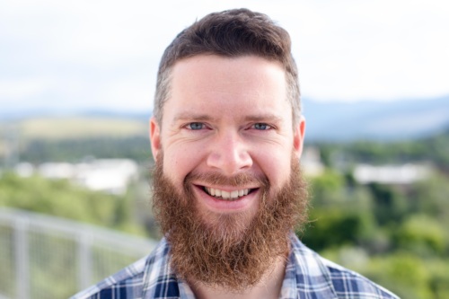 Headshot of Ben Lyon, a white man with a long beard in front of scenic mountains