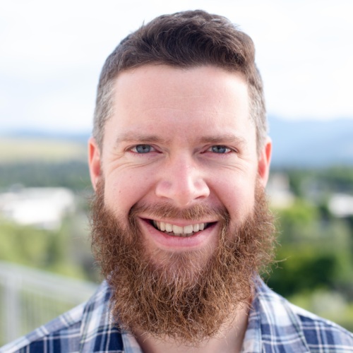 Headshot of Ben Lyon, a white man with a long beard in front of scenic mountains