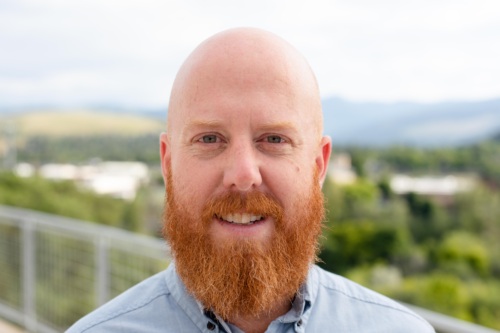 Headshot of Ryan Smither, a white bald man with a long beard standing in front of scenic mountains.