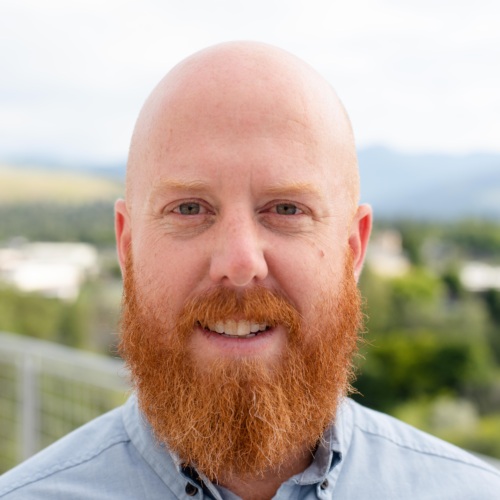 Headshot of Ryan Smither, a white bald man with a long beard standing in front of scenic mountains.