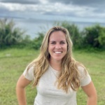 Photo of Sara Hunold, a white woman with long blonde hair standing in the grass in front of the beach