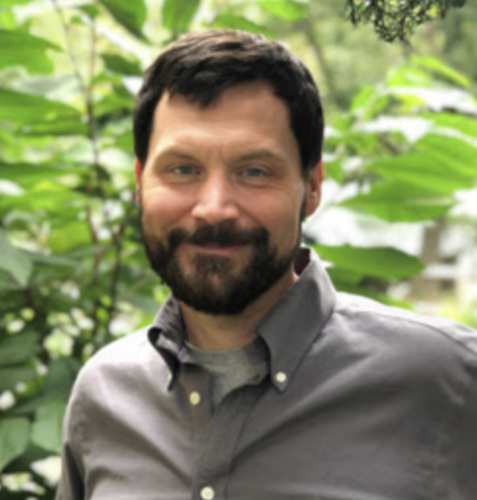 Picture of Alex Ireland, a white middle aged man with brown hair and a beard standing in front of trees.