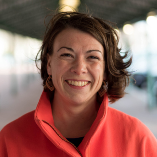Headshot image of a white woman with short bobbed and windswept brunette hair in coral jacket.